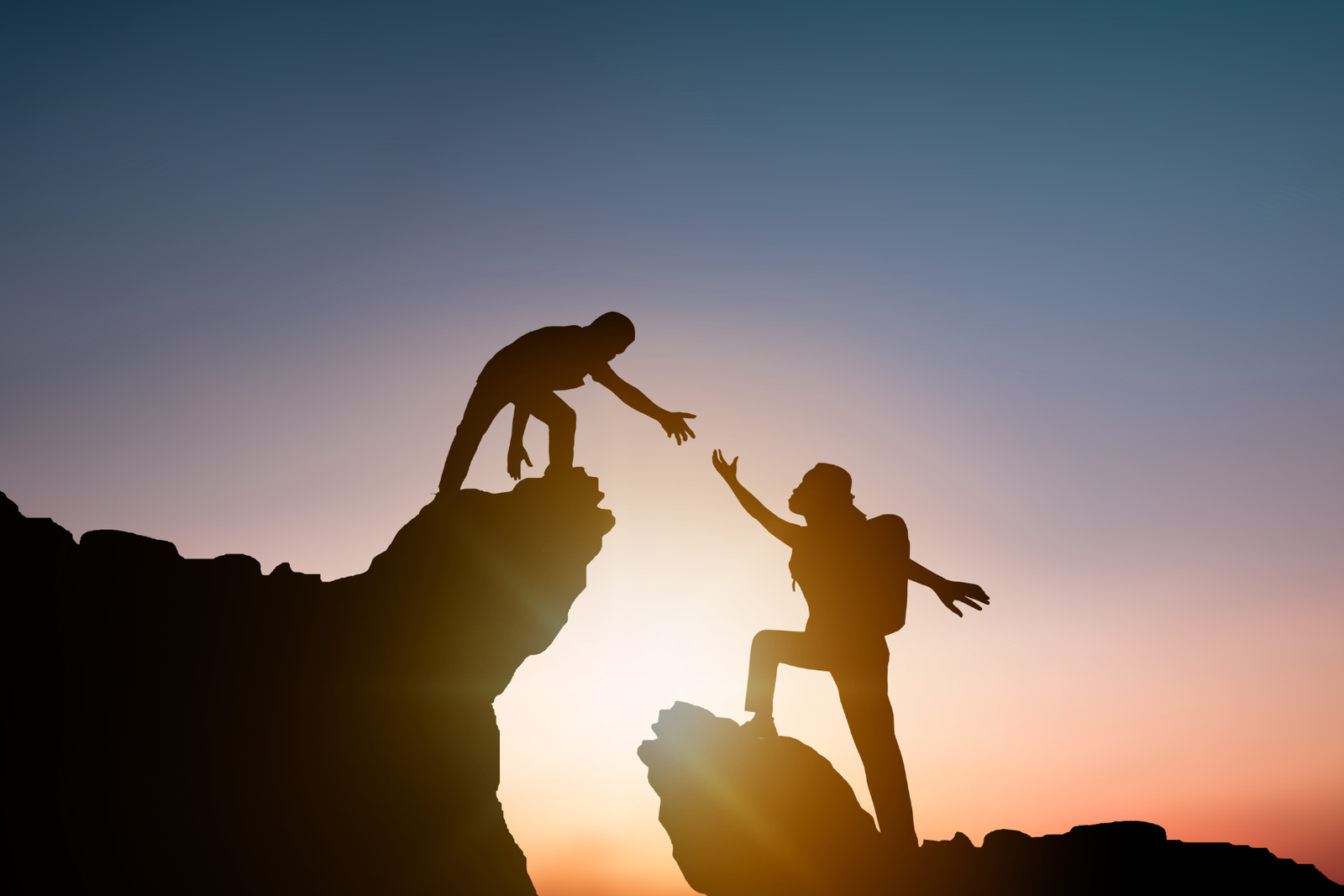 Silhouette of two hikers at sunrise or sunset, with one person reaching down to help another climb up a steep rock, symbolizing teamwork, support, and achievement.
